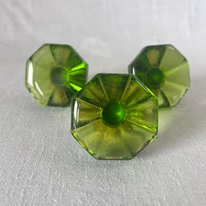 May include: Three green glass cabinet knobs with an octagonal shape. Each knob has a central, rounded detail and radiating lines. The knobs are a vibrant green color and appear to be made of glass.