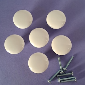 May include: Six off-white, round cabinet knobs with a glossy finish are arranged on a purple surface.  Five knobs are positioned in a circular pattern, with the sixth knob slightly offset.  Four silver screws are also included.
