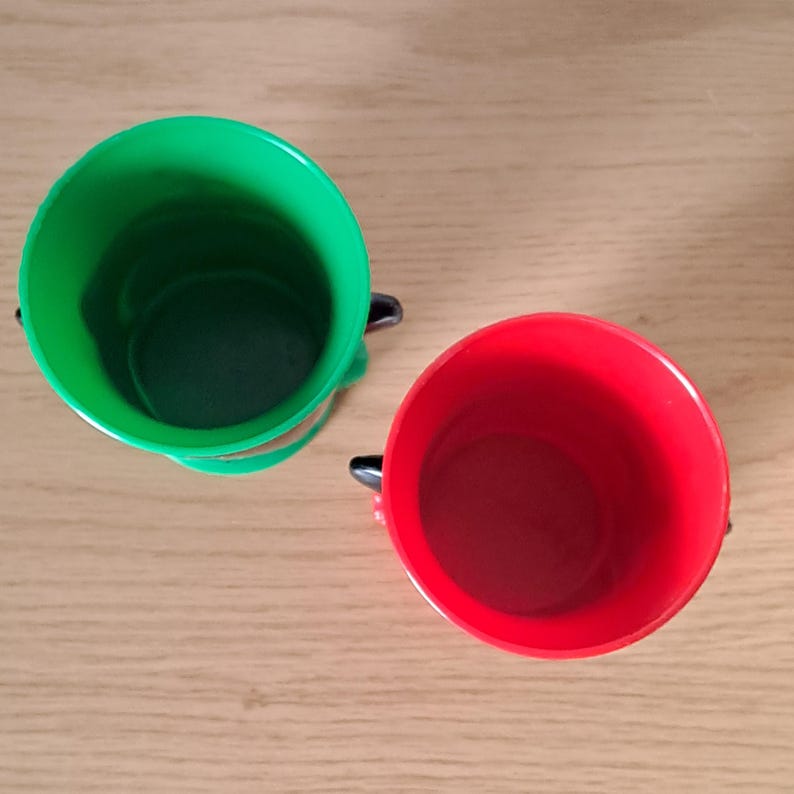 May include: Two plastic buckets, one green and one red, are shown from a top-down perspective. Each bucket has a black handle and a rounded shape. The buckets are sitting on a light brown wooden surface.