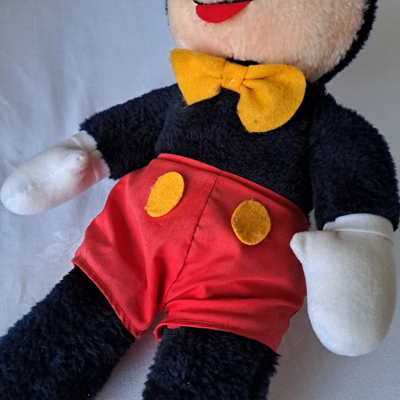 Puede incluir: Un juguete de peluche de Mickey Mouse con pelaje negro, pantalones cortos rojos con botones amarillos y una pajarita amarilla.