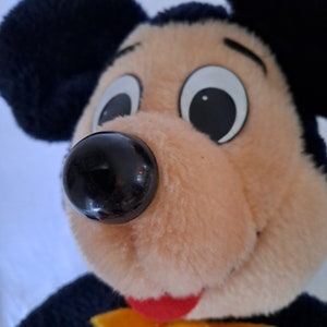 Puede incluir: Primer plano de un juguete de peluche de Mickey Mouse con orejas negras, nariz negra y grandes ojos blancos. El juguete tiene una cara marr&oacute;n claro y una lengua roja.