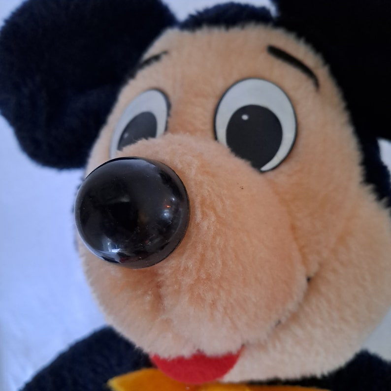 Puede incluir: Primer plano de un juguete de peluche de Mickey Mouse con orejas negras, nariz negra y grandes ojos blancos. El juguete tiene una cara marr&oacute;n claro y una lengua roja.