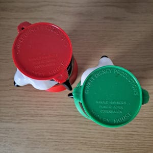 May include: Two vintage Mickey Mouse mugs. One mug has a red lid, the other has a green lid. Both mugs have the text "Walt Disney Productions" and "Mickey Mouse" printed on them.