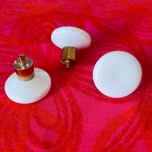 May include: Three circular cabinet knobs with smooth white tops and metallic gold-toned cylindrical bases. Each knob features a small threaded screw post extending from the center of the base, designed for attachment to furniture or cabinetry doors.
