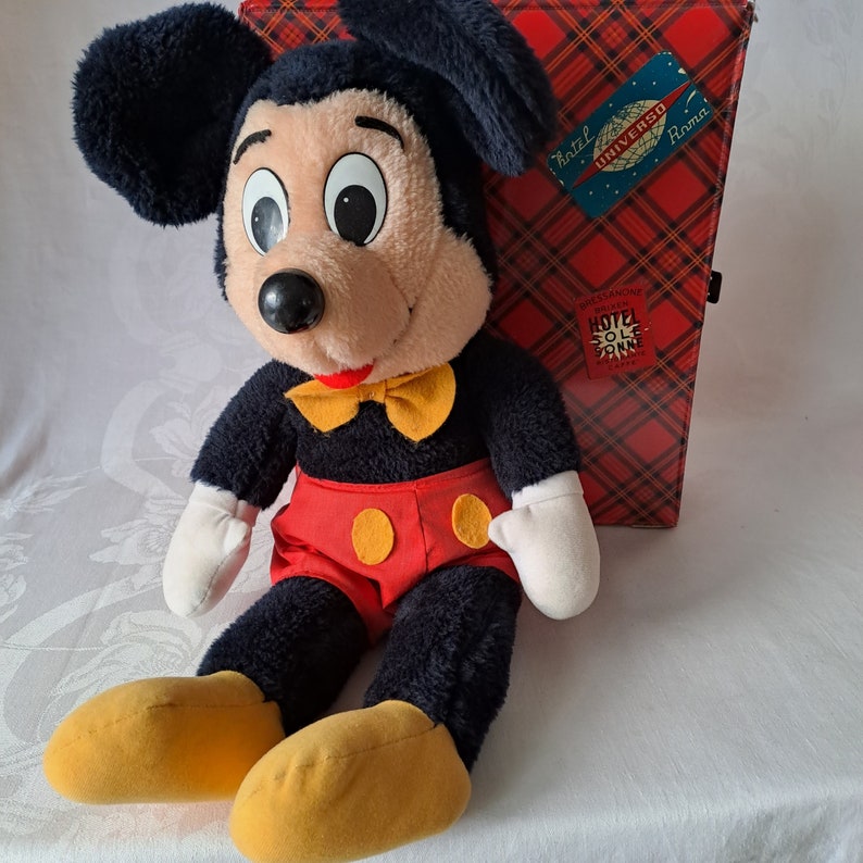 Puede incluir: Un juguete de peluche Mickey Mouse negro y rojo con una pajarita amarilla sentado junto a una maleta de tela a cuadros rojos y negros con el texto "Hotel Universo Roma" y "Hotel Sole Rimini" en ella.