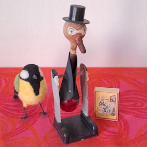 May include: A whimsical collection of vintage toys and collectibles. Includes a felt bird, a wooden bird figure wearing a top hat and suit, and a small matchbox with a vintage illustration. The bird figure has a red glass bulb.