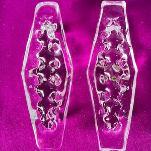 Op de afbeelding: Twee transparante glazen decoratieve objecten met een getextureerd, golvend ontwerp. De objecten zijn langwerpig diamantvormig en hebben een licht gematteerde uitstraling.