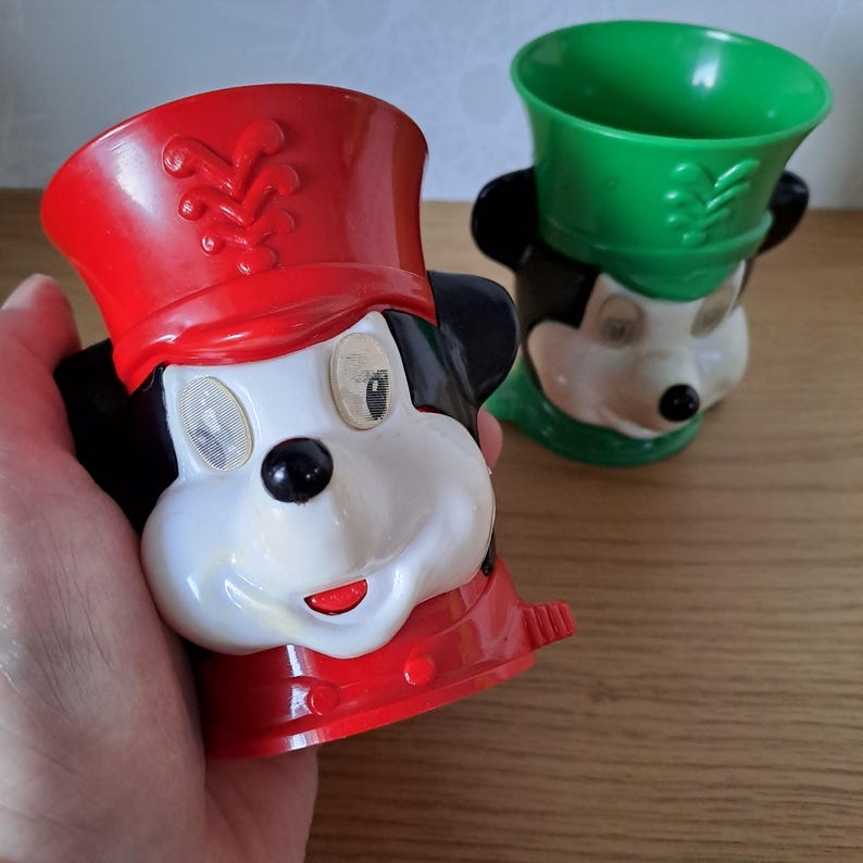 May include: Two vintage Mickey Mouse character cups. One cup is held in a hand and is red, black, and white. The other cup is green, black, and white. Both cups are in the shape of Mickey Mouse's head and feature a hat.