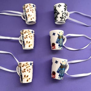 Könnte beinhalten: Sechs kleine, weiße Keramikbecher mit Henkeln und dekorativen Mustern. Die Becher weisen florale, Eulen- und geometrische Designs in Blau, Braun und Schwarz auf. Jeder Becher hat ein weißes Band.