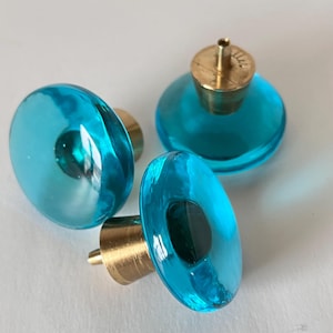 Può includere: Tre pomelli rotondi per mobili con parte superiore in vetro turchese traslucido e superfici lisce e bombate. Ogni pomello è fissato a una base cilindrica in metallo color ottone con un piccolo bullone filettato che sporge dal centro.