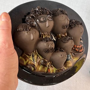 Könnte beinhalten: Eine Keramik-Wandplatte mit einer Gruppe von sechs braunen Figuren mit übertriebenen Merkmalen, darunter große Augen und Nasen. Die Figuren sind in einem Halbkreis angeordnet, wobei die größte Figur in der Mitte steht. Die Platte hat einen schwarzen Hintergrund und eine glänzende Oberfläche.