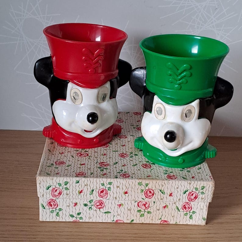 May include: Two vintage Mickey Mouse character cups. One cup is red, the other is green. Both cups have black ears and a white face. The cups are sitting on a floral patterned box.