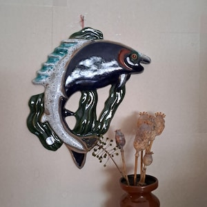 May include: A ceramic fish wall hanging in navy blue, white, and green. The fish has a glossy finish and is mounted on a neutral wall. The fish is surrounded by green aquatic plants. The artwork is a decorative piece.