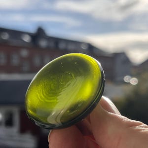 Può includere: Un oggetto ovale in vetro verde oliva liscio. Il vetro ha una finitura lucida e sono visibili vortici interni. L'oggetto è tenuto contro uno sfondo sfocato all'aperto, con edifici e cielo visibili.