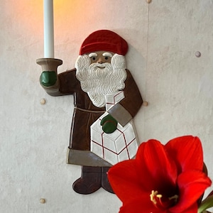May include: Wooden Santa Claus wall decoration holding a lit candle and a wrapped gift. The Santa figure has a red hat, white beard, and brown coat. A vibrant red flower is in the foreground. The candle is white and the flame is yellow.