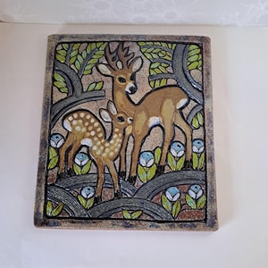 May include: Rectangular ceramic tile featuring a deer and fawn in a forest scene. The deer are brown with detailed antlers, and the fawn is spotted. The background includes stylized trees, leaves, and blue and white flowers. The tile has a textured, earthy color palette.