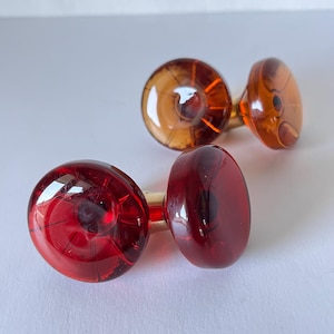 May include: A pair of cufflinks with a red and amber glass design. The cufflinks have a rounded shape with a gold-colored metal post. The glass has a transparent quality, allowing light to pass through.