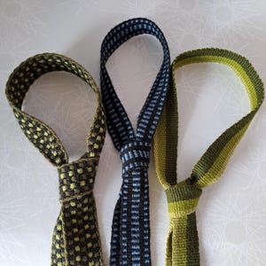 May include: Three woven neckties in shades of green, blue, and black. The ties are knotted at the bottom and looped at the top. The patterns include a checkered design and striped pattern.