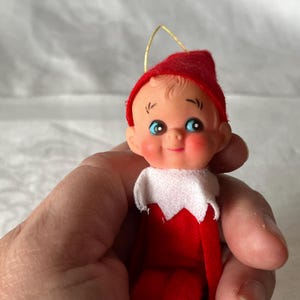 Peut inclure: Ornement de Noël vintage représentant un elfe. Il porte un chapeau et une tenue en feutre rouge, un col blanc et un visage souriant avec des yeux bleus et des joues roses. Une ficelle dorée est attachée pour l'accrocher.