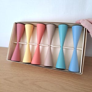 May include: A set of six hourglass-shaped candle holders in a box. The candle holders are in pastel shades of pink, yellow, coral, beige, teal, and blue. The box is beige and the candle holders are made of wood.