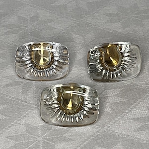 Op de afbeelding: Drie transparante glazen knoppen met een goudkleurig ontwerp in het midden. De knoppen hebben een zonnestralenpatroon rond het middelste ontwerp.