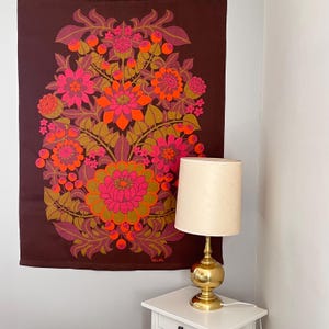 May include: A brown floral tapestry with pink, orange, and green flowers hangs on a wall. The tapestry has a large, stylized flower design. A gold-coloured lamp with a cream-coloured shade sits on a white bedside table.