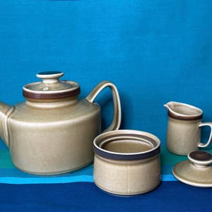 Könnte beinhalten: Ein braunes und beiges Keramik-Set aus Teekanne, Zuckerdose und Milchkännchen. Die Teekanne hat einen braunen Deckel und einen braunen Streifen um den oberen Rand. Die Zuckerdose hat einen braunen Deckel und einen braunen Streifen um den oberen Rand. Das Milchkännchen hat einen braunen Streifen um den oberen Rand.