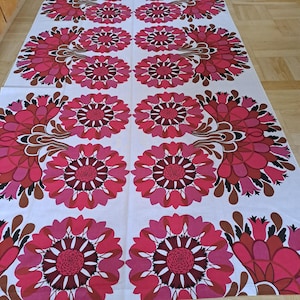 May include: A white tablecloth with a repeating floral pattern in shades of pink, red, and brown. The flowers have a stylized, retro design, with a central dark red circle surrounded by petals.