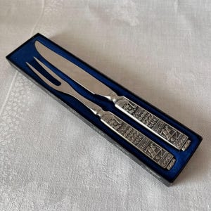 May include: A carving set comprising a knife and fork, displayed in a black box with a blue lining. The silver-coloured utensils have ornate handles with engraved designs. The knife has a curved blade, and the fork has two prongs.