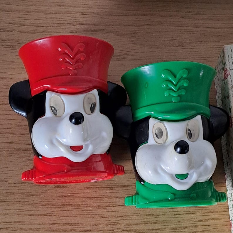 May include: Two vintage Mickey Mouse character-shaped cups. One cup is red, and the other is green. Both cups have black ears, white faces, and a red or green hat with a decorative design. The cups are made of plastic and are likely from the 1980s or 1990s.
