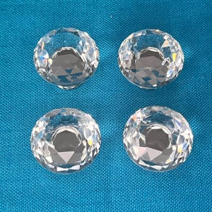 Può includere: Quattro pomelli di cristallo trasparente e sfaccettato con un design circolare. I pomelli sono disposti su una superficie di tessuto blu. I pomelli di cristallo hanno una qualità riflettente, che cattura e rifrange la luce.