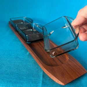 Puede incluir: Una bandeja de madera con tres cuencos cuadrados de vidrio. Los cuencos de vidrio son de color gris ahumado y tienen asas pequeñas. La bandeja es de color marrón oscuro y tiene forma curva.