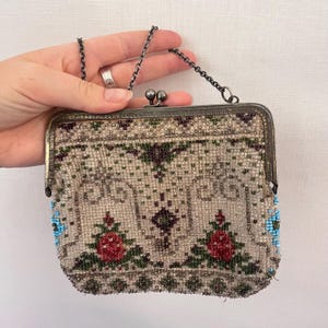 Op de afbeelding: Vintage kralen tas met een zilverkleurig metalen frame en ketting handvat. De tas heeft een bloemenontwerp met rode rozen, groene bladeren en blauwe accenten. Het kralenpatroon is ingewikkeld en gedetailleerd en toont een klassieke, elegante stijl.