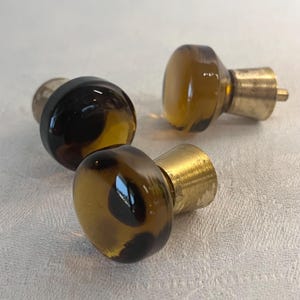 Op de afbeelding: Drie bruine glazen kastknoppen met messing bases. De knoppen zijn rond en hebben een licht getextureerd oppervlak.