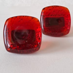May include: Two square, red glass cabinet knobs with rounded corners. The knobs have a textured surface and a dark rectangular shape in the centre. The glass is translucent, allowing light to pass through.