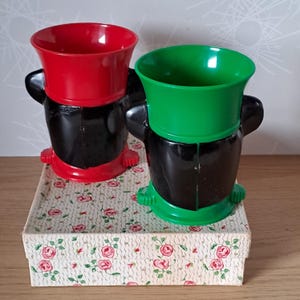 May include: Two vintage plastic novelty cups, one red and one green, with black bases and handles. They sit on a floral patterned box. The cups have a unique, cartoonish design.