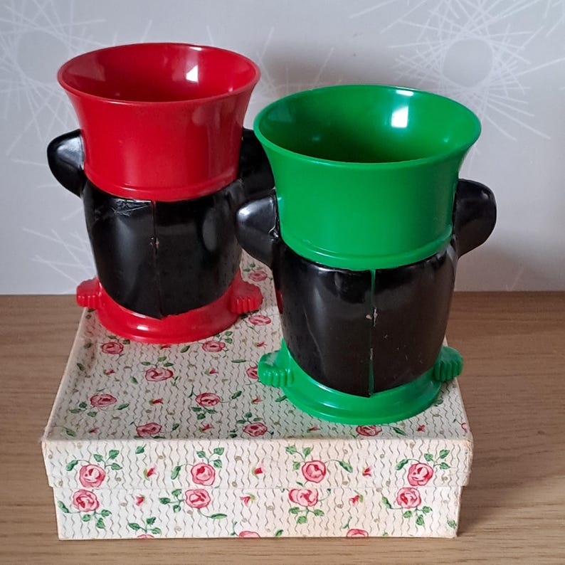 May include: Two vintage plastic novelty cups, one red and one green, with black bases and handles. They sit on a floral patterned box. The cups have a unique, cartoonish design.