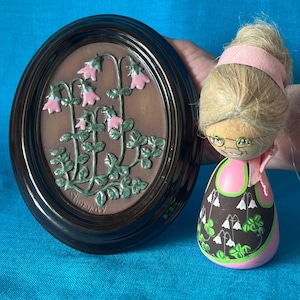 May include: An oval, dark brown framed relief sculpture featuring pink flowers and green leaves. Beside it is a wooden doll with blonde hair, glasses, and a pink and black dress. The doll has a pink headband and scarf.
