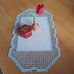 Peut inclure: Nappe rectangulaire bleu clair avec une bordure en crochet. Une boîte rouge avec des motifs floraux et un petit vase contenant une tulipe rouge sont sur la nappe. La nappe est sur une table en bois.