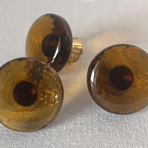 Op de afbeelding: Drie bruine glazen kastknoppen met een getextureerd oppervlak en een donkerbruin centrum. De knoppen hebben een gouden metalen bevestiging aan de achterkant.