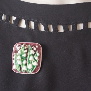 May include: A square ceramic brooch with a raised design of white lily of the valley flowers with green leaves on a burgundy background.