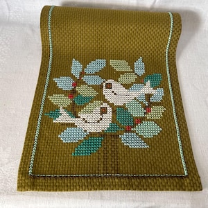 Puede incluir: Camino de mesa de punto de cruz verde oliva con un diseño de dos pájaros blancos posados en una rama frondosa con detalles azules, verdes y rojos. Los bordes están ribeteados con una puntada azul claro.