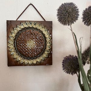 Könnte beinhalten: Eine braune und cremefarbene Keramikfliese mit einem Sonnenblumenmuster. Die Fliese hat eine erhabene, strukturierte Oberfläche und ist von einem braunen Rahmen umgeben.