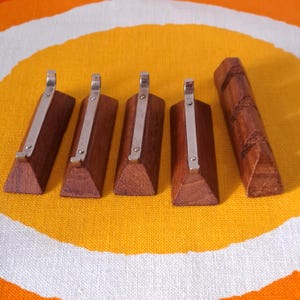 Pode incluir: Um conjunto de cinco argolas de guardanapo de madeira. Quatro têm uma forma retangular com um clipe de metal prateado, e uma tem um design esculpido. As argolas de guardanapo são de cor castanho escuro e estão sobre uma superfície estampada em amarelo e laranja.