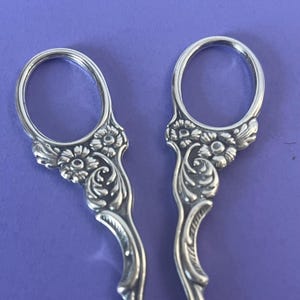 Puede incluir: Tijeras de bordar plateadas con detalles florales y de volutas. Los mangos presentan lazos abiertos para los dedos y adornos florales decorativos. Las tijeras están sobre un fondo morado.
