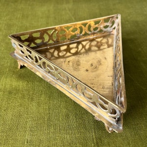 Può includere: Un vassoio triangolare in ottone con un intricato design traforato attorno al bordo. Il vassoio ha tre gambe ed è realizzato in un metallo lucido color oro.