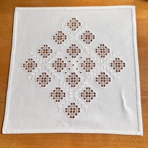 Peut inclure: Nappe carrée en lin blanc avec un motif géométrique brodé. Le motif présente une forme de losange central avec de plus petits losanges et des motifs en forme de feuilles. La broderie est blanche, créant un effet texturé sur le fond lisse.
