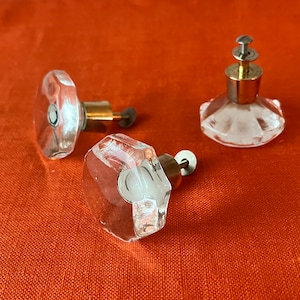 Op de afbeelding: Drie heldere glazen kastknoppen met messing beslag. De knoppen hebben een facetgeslepen ontwerp en zijn in verschillende vormen.