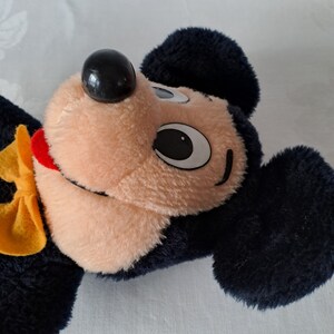 Puede incluir: Un juguete de peluche de Mickey Mouse con orejas negras, una nariz negra y una pajarita amarilla. El juguete tiene una cara marr&oacute;n claro con ojos negros y una boca roja.