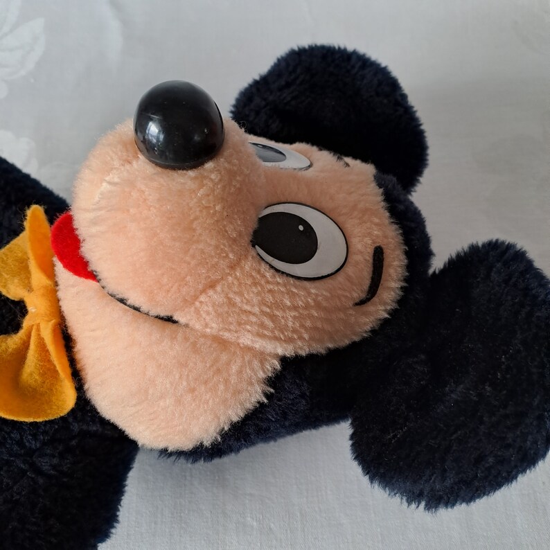 Puede incluir: Un juguete de peluche de Mickey Mouse con orejas negras, una nariz negra y una pajarita amarilla. El juguete tiene una cara marr&oacute;n claro con ojos negros y una boca roja.